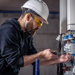 A male electrician works in a switchboard with an electrical connecting cable, connects the equipment with tools.